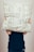 A person is holding a pillow covered with newspaper print patterns in front of their face. The person is wearing a dark blue top and is visible from the shoulders down. The background is plain and light-colored.