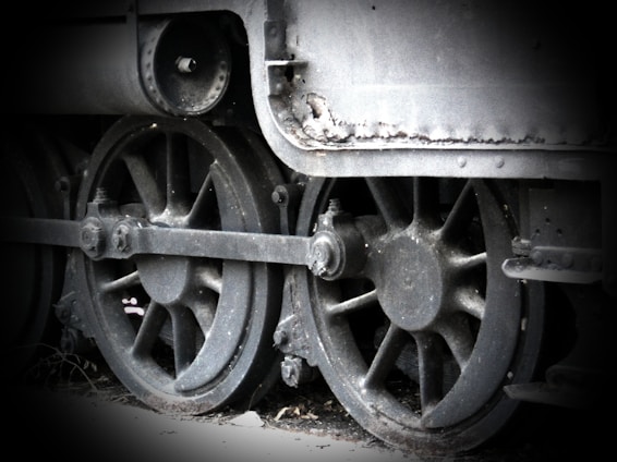 A black and white image featuring the side view of an old industrial train's large metal wheels. The wheels are connected by rods and exhibit signs of wear and rust, adding a vintage or historic feel.