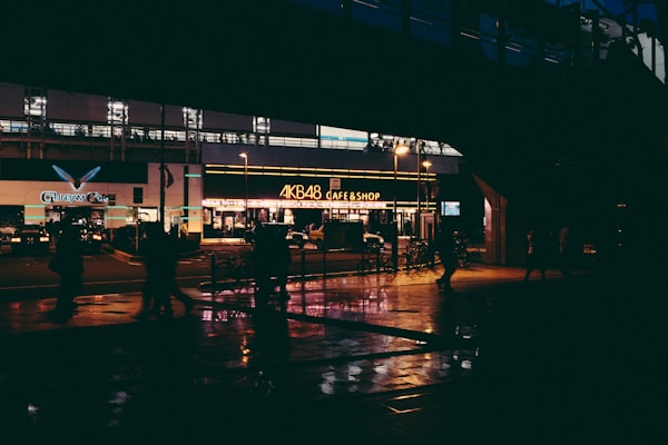 A nighttime city street scene featuring a brightly lit cafe and shop with glowing signage. The atmosphere is enhanced by streetlights and reflections on the wet pavement. Several silhouetted figures can be seen walking along the sidewalk, creating an urban nightlife vibe.