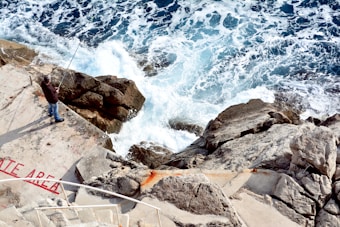 A person is fishing on a rocky shoreline with waves crashing against the rocks. The angler stands on a concrete area labeled 'PRIVATE AREA' in red paint. Stairs lead down to the platform, and the sea is a mix of deep blue and foamy white waves.