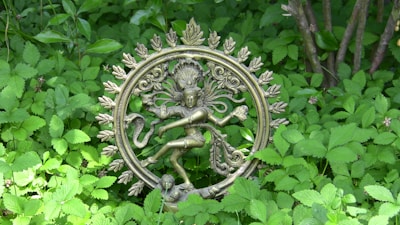 A bronze statue of Nataraja, the Hindu deity Shiva in his cosmic dance form, is surrounded by lush green foliage. The intricate detailing of the sculpture features Shiva dancing within a circular arch of flames, signifying the cosmic cycles of creation and destruction.