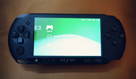 A handheld gaming console with a screen displaying icons and options for games and saved data. The device has typical gaming buttons and directional pads on either side of the screen.
