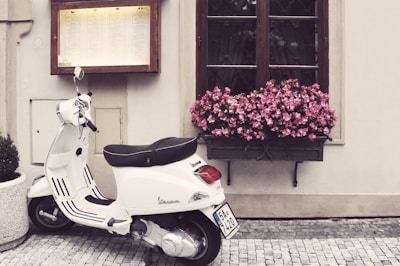 A white scooter is parked on a cobblestone street next to a beige wall. Above the scooter, there is a large wooden-framed window with a box filled with vibrant pink flowers. Next to the window, a menu is displayed in a wooden case.