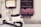 A white scooter is parked on a cobblestone street next to a beige wall. Above the scooter, there is a large wooden-framed window with a box filled with vibrant pink flowers. Next to the window, a menu is displayed in a wooden case.