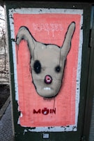A poster with a stylized, abstract face featuring large, dark eyes and prominent ears. The background is a salmon pink color, with the words 'KASSEL' and 'MOIN' appearing at the top and bottom. The face is outlined in black with drips of paint emphasizing a raw, street art aesthetic.