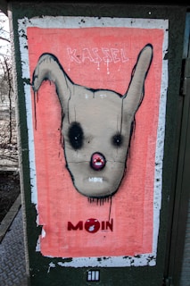 A poster with a stylized, abstract face featuring large, dark eyes and prominent ears. The background is a salmon pink color, with the words 'KASSEL' and 'MOIN' appearing at the top and bottom. The face is outlined in black with drips of paint emphasizing a raw, street art aesthetic.