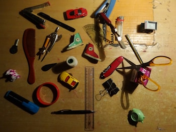 A collection of various household items scattered on a wooden surface. Items include a red comb, a compass, toy cars, a ruler, scissors, a folding ruler, a lighter, a stapler, a roll of tape, a stapler remover, and a highlighter. Most items are placed randomly in different orientations.