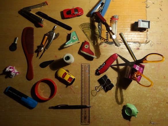 A collection of various household items scattered on a wooden surface. Items include a red comb, a compass, toy cars, a ruler, scissors, a folding ruler, a lighter, a stapler, a roll of tape, a stapler remover, and a highlighter. Most items are placed randomly in different orientations.