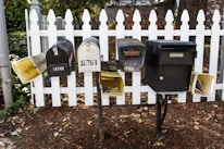 A group of five mailboxes is mounted on black metal posts in front of a white picket fence. There are various designs and sizes, some vintage-looking, with addresses visible on each. Fallen leaves are scattered on the ground around the base of the fence.