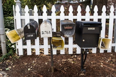 A group of five mailboxes is mounted on black metal posts in front of a white picket fence. There are various designs and sizes, some vintage-looking, with addresses visible on each. Fallen leaves are scattered on the ground around the base of the fence.