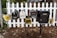 A group of five mailboxes is mounted on black metal posts in front of a white picket fence. There are various designs and sizes, some vintage-looking, with addresses visible on each. Fallen leaves are scattered on the ground around the base of the fence.