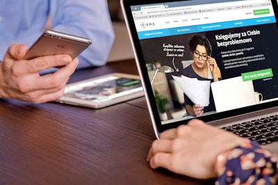 A person is holding a smartphone near a laptop displaying a website. The laptop screen shows a woman holding papers and talking on the phone, along with text in Polish. A tablet is also visible on the table.