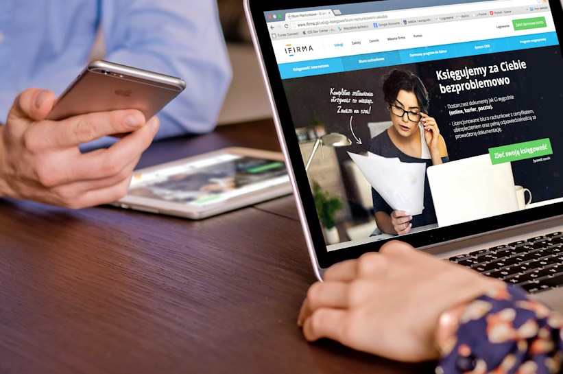 A person is holding a smartphone near a laptop displaying a website. The laptop screen shows a woman holding papers and talking on the phone, along with text in Polish. A tablet is also visible on the table.