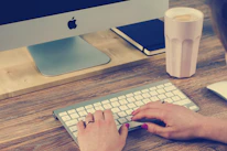 Close-up of hands typing on a wireless keyboard with a blurred coffee cup nearby.