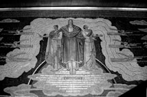 A detailed mural depicting three central human figures dressed in draped garments, possibly indicating classical or historical influences. The central figure raises an arm, and they are flanked by two others who are gesturing outward. Surrounding them are clouds and what appears to be winged human forms or mythical creatures. The word 'THOUGHT' is prominently displayed beneath the central figure's feet.