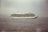A large cruise ship sails on the calm sea, with a slight haze in the sky and a distant coastline visible on the left. The ship is detailed with multiple decks, and lifeboats are visible on the side.