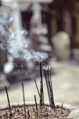 A bundle of incense sticks burning in an ash-filled container, releasing thin spirals of smoke into the air. The background is blurred, creating a soft focus effect.