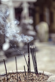 A bundle of incense sticks burning in an ash-filled container, releasing thin spirals of smoke into the air. The background is blurred, creating a soft focus effect.