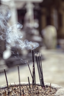 A bundle of incense sticks burning in an ash-filled container, releasing thin spirals of smoke into the air. The background is blurred, creating a soft focus effect.
