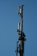 A tall communication tower equipped with multiple antennas and equipment, set against a clear blue sky. The structure appears metallic with various protruding components and cables.
