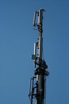 A tall communication tower equipped with multiple antennas and equipment, set against a clear blue sky. The structure appears metallic with various protruding components and cables.