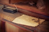 A dusty wooden surface holds an old, yellowed newspaper with visible text and a photograph. Adjacent to the newspaper is an aged, portable cassette player and a few indistinct objects, suggesting an abandoned or nostalgic setting with a sense of clutter and neglect.