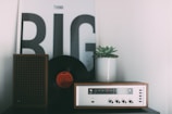 A vintage radio sits on a surface alongside a vinyl record and a small brown speaker. A potted succulent plant is also present. In the background, there is a large poster with the words 'Think Big' in bold black letters.