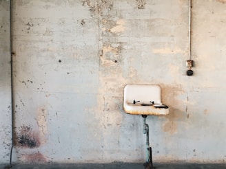 A worn and rusted utility sink mounted on an aged and cracked concrete wall. The surroundings appear to be part of an old industrial or abandoned area, featuring exposed pipes and weathered, peeling paint.