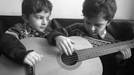 Two children are wearing patterned sweaters and sitting closely together, focused on playing a guitar. Their expressions appear concentrated as one child carefully plucks the strings while the other watches attentively.