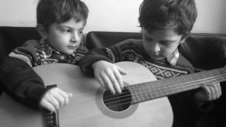 Two children are wearing patterned sweaters and sitting closely together, focused on playing a guitar. Their expressions appear concentrated as one child carefully plucks the strings while the other watches attentively.