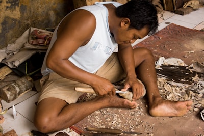 A person is engaged in woodworking, sitting cross-legged on the floor surrounded by wood shavings. They are carving a piece of wood using a chisel and a mallet. There are various tools and materials scattered around, including a knife and a block of wood.