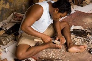A person is engaged in woodworking, sitting cross-legged on the floor surrounded by wood shavings. They are carving a piece of wood using a chisel and a mallet. There are various tools and materials scattered around, including a knife and a block of wood.