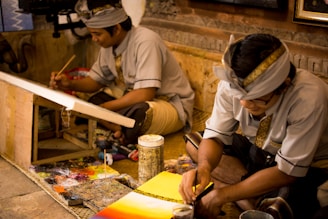 Two individuals are wearing traditional attire and are engaged in a painting activity. They are seated on a woven mat, surrounded by art supplies including paintbrushes and a wooden easel. The environment appears to be an artistic workspace with a warm and focused atmosphere.