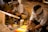 Two individuals are wearing traditional attire and are engaged in a painting activity. They are seated on a woven mat, surrounded by art supplies including paintbrushes and a wooden easel. The environment appears to be an artistic workspace with a warm and focused atmosphere.