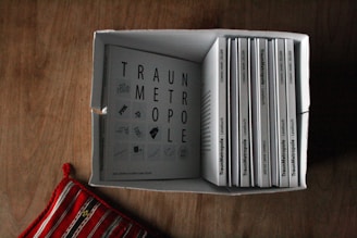 A box filled with multiple copies of a book titled 'Traummetropole'. The books are neatly arranged side by side with one lying flat on top. A patterned red and white textile is placed next to the box on a wooden surface.