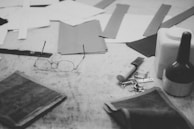 A collection of crafting tools and materials on a work surface. Items include eyeglasses, scissors, pieces of fabric, a glue bottle, and some keys. The scene has a monochromatic and vintage feel, with a focus on the artisan nature of the objects.