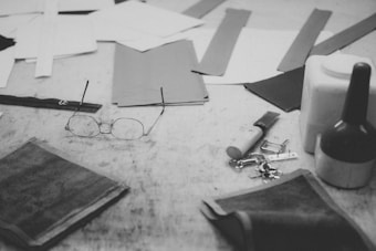A collection of crafting tools and materials on a work surface. Items include eyeglasses, scissors, pieces of fabric, a glue bottle, and some keys. The scene has a monochromatic and vintage feel, with a focus on the artisan nature of the objects.