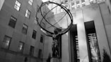 A large, striking sculpture depicting a muscular figure holding an armillary sphere on their shoulders. The sculpture is situated in an urban setting, with towering buildings surrounding it. The scene conveys a sense of grandeur and strength.