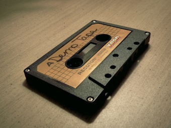 A vintage audio cassette tape with a handwritten label reading 'Demo Tape' is placed on a wooden surface. The cassette has a grid design with numbers indicating minutes, and the brand 'YR90' visible near the corner.