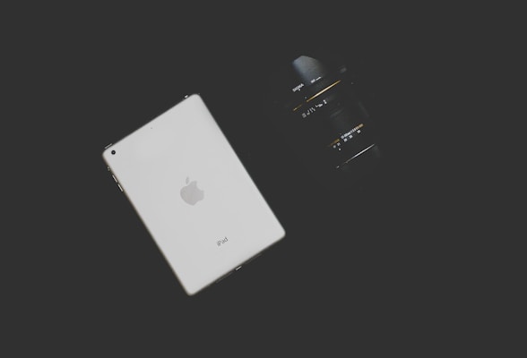 A white tablet with an Apple logo is placed on a dark background alongside a black camera lens. The tablet is positioned on the left, and the lens on the right is partially in shadow.