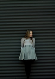 A woman stands against a dark, textured wall with horizontal stripes. She is wearing a light-colored blouse and a gray skirt with buttons, her hands tucked into the pockets on either side. Her long brown hair falls loosely over her shoulders, and she gazes slightly downward with a peaceful expression.