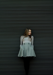 A stylish woman in a soft beige shirt and muted earth tone pants, standing against a textured wall with a relaxed, poised expression