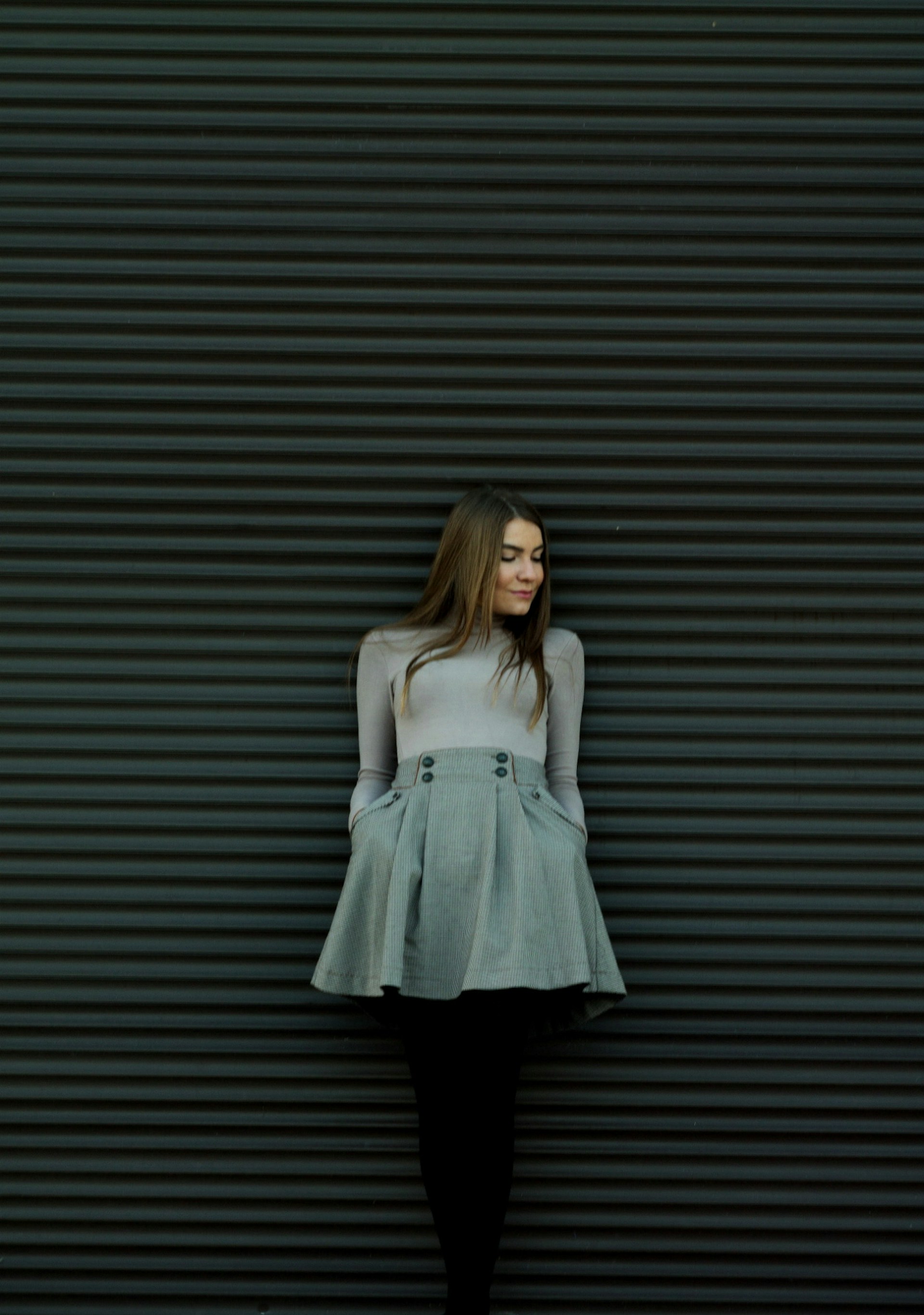 A relaxed woman in a flowing white blouse and tailored pants standing against a pale stone wall, bathed in gentle natural light.