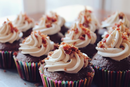 A row of chocolate cupcakes topped with creamy white frosting and small pieces of what appears to be bacon bits. The cupcakes are in colorful striped paper liners, creating a visually appealing contrast with the frosting.