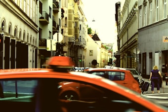 A bustling urban street scene with historic architecture lining both sides. The buildings are mainly composed of earthy tones, featuring balconies and arched windows. Several cars, including a prominent red vehicle, are in motion, calling attention to the city's active atmosphere. A person is walking along the sidewalk, pulling a suitcase.