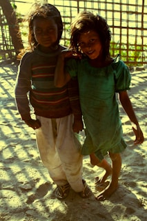 Two children stand on a sandy ground with sunlight casting shadows through a bamboo structure in the background. One child wears a green dress while the other is in a colorful sweater and white pants. Both children have dark, wavy hair and are smiling.