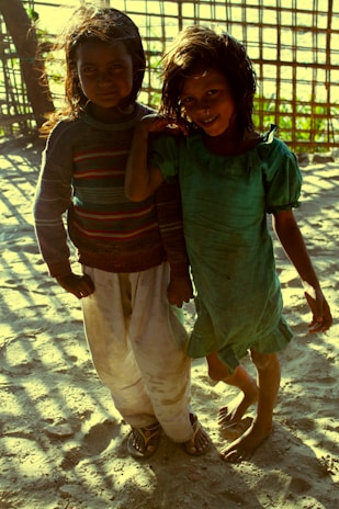 Two children stand on a sandy ground with sunlight casting shadows through a bamboo structure in the background. One child wears a green dress while the other is in a colorful sweater and white pants. Both children have dark, wavy hair and are smiling.