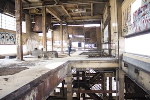 An abandoned industrial building interior with large windows partially covered in graffiti. The structure is visibly worn, featuring exposed beams, rusted metal elements, and peeling paint. A person is standing near the center, examining the space.