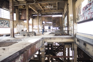 An abandoned industrial building interior with large windows partially covered in graffiti. The structure is visibly worn, featuring exposed beams, rusted metal elements, and peeling paint. A person is standing near the center, examining the space.
