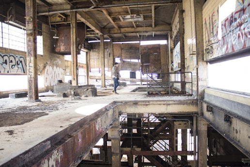 An abandoned industrial building interior with large windows partially covered in graffiti. The structure is visibly worn, featuring exposed beams, rusted metal elements, and peeling paint. A person is standing near the center, examining the space.